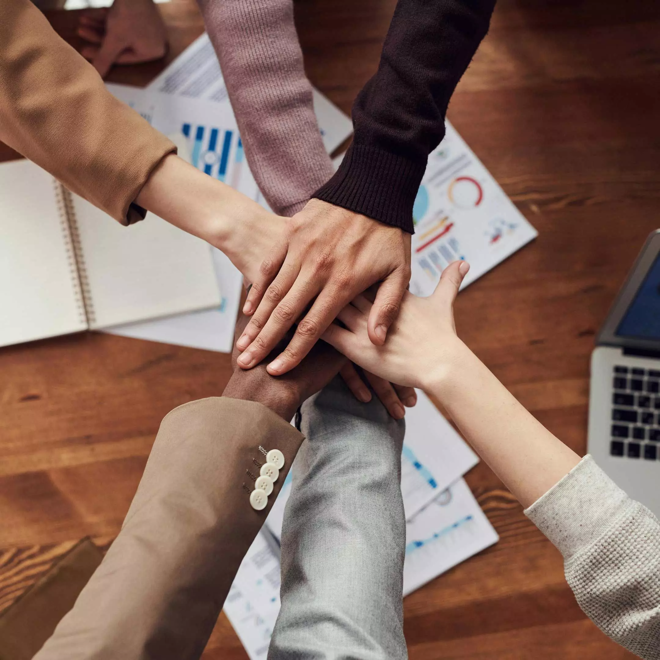 A team of professionals in business attire joins hands in unity over printed financial reports, symbolizing collaboration and shared success.